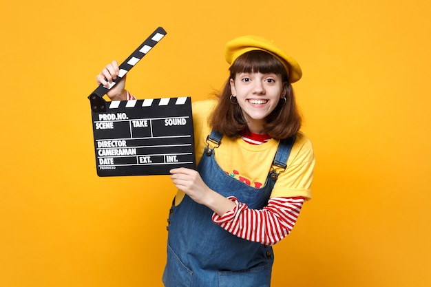 Funny girl teenager in french beret, denim sundress holding classic black film making clapperboard isolated on yellow wall background. People sincere emotions lifestyle concept. Mock up copy space.