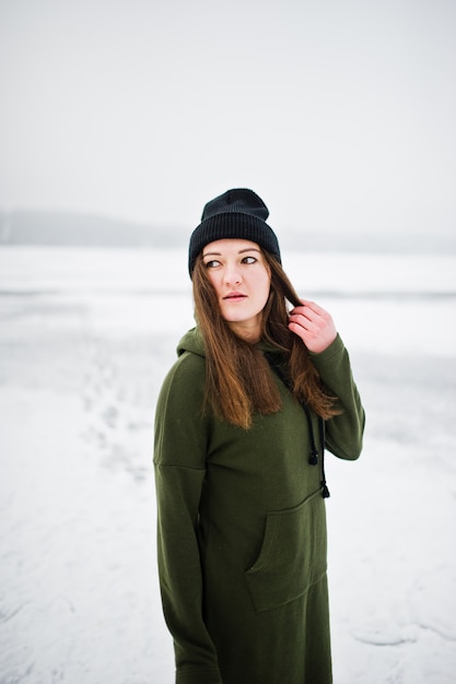 Funny girl wear on long green sweatshirt, jeans and black headwear, at frozen lake in winter day.