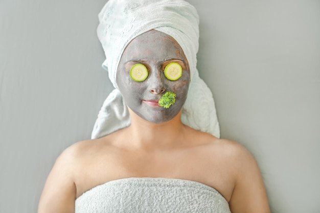 Funny girl with cosmetic mask made of gray clay and pieces of vegetables on face, her hair wrapped i