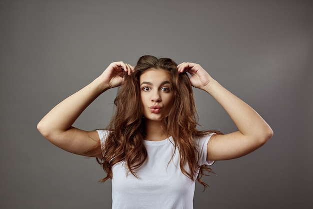 Funny girl with long brown hair dressed in a white t-shirt holds her hands on her head in the studio on the gray background .