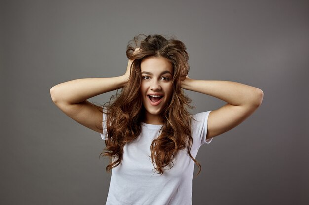 Funny girl with long brown hair dressed in a white t-shirt holds her hands on her head in the studio on the gray background .