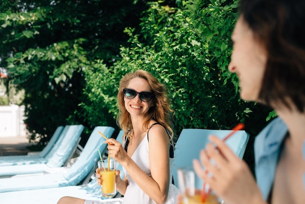 Ragazze divertenti parlano di bere succo di frutta e rilassarsi seduti su chaise longue vicino alla piscina dell'hotel o della villa vacanze estive e riposo dopo il lavoro in hotel per tutte le stagioni disintossicazione del corpo e spa viaggi