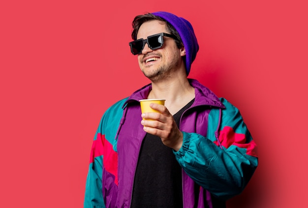 Photo funny guy in 80s style jacket and sunglasses holds paper cup on red backgorund