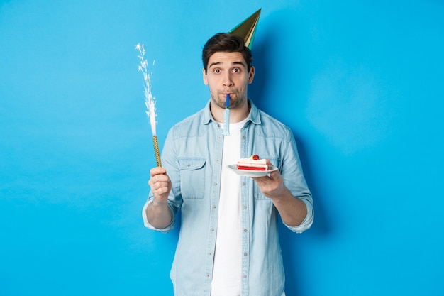 Funny guy celebrating birthday, holding b-day cake, firework and wearing party hat, standing over blue background.