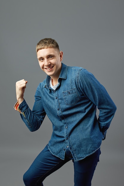 Funny guy dressed in a jeans shirt shows his power with his hand in the studio on the gray background .