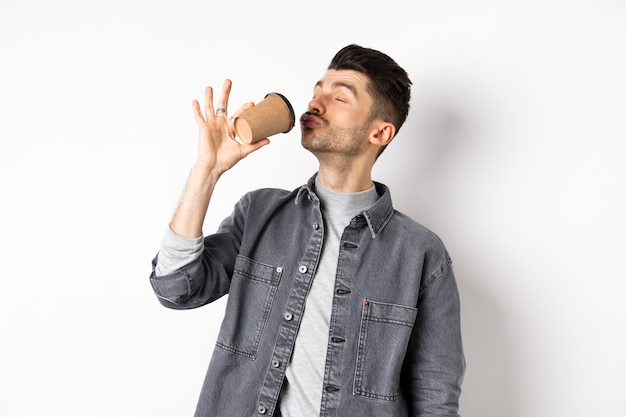 白い背景の上に立って、満足のいくカフェから持ち帰り用の飲み物をすすりながら、カップからコーヒーを飲むことを楽しんでいる面白い男。