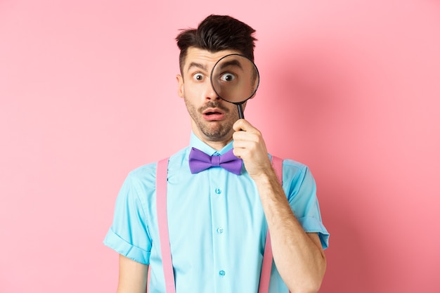 Funny guy look through magnifying glass with surprised face, seeing something interesting, standing on pink background.