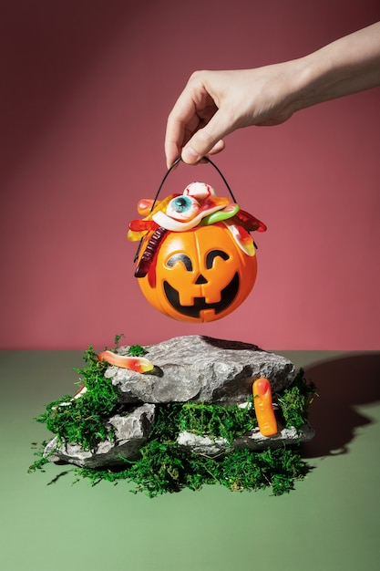 Funny Halloween scenery. Trick or treat. Pumpkin Jack filled with various creepy sweets stands on stones and moss. Woman hand holds basket