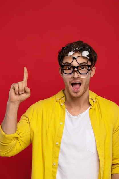 funny handsome man with open mouth in yellow shirt with many eyeglasses on face showing idea gesture isolated on red  background