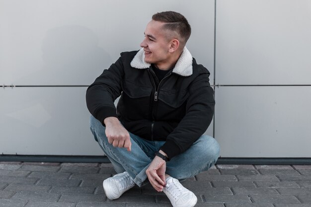 Funny handsome young man with a fashionable hairstyle in a stylish jacket in vintage jeans in sneakers sits and smiles near the modern building on a bright spring day