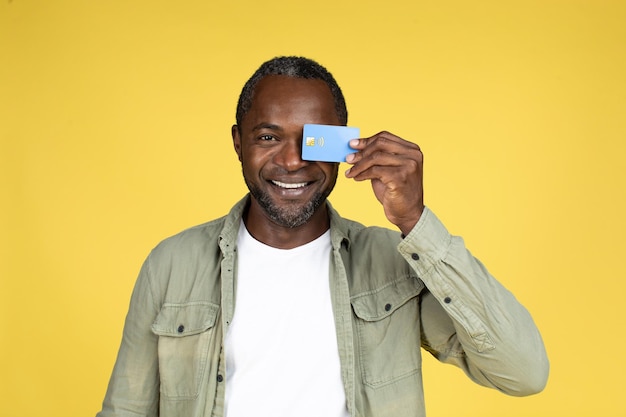 Funny happy adult black guy in casual puts credit card to eye rejoices to great online shopping have fun