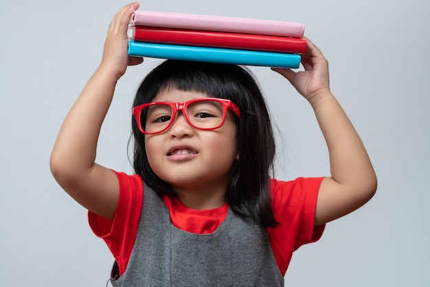 頭に緑の本を持って赤い眼鏡をかけている面白くて幸せなアジアの小さな就学前の女の子