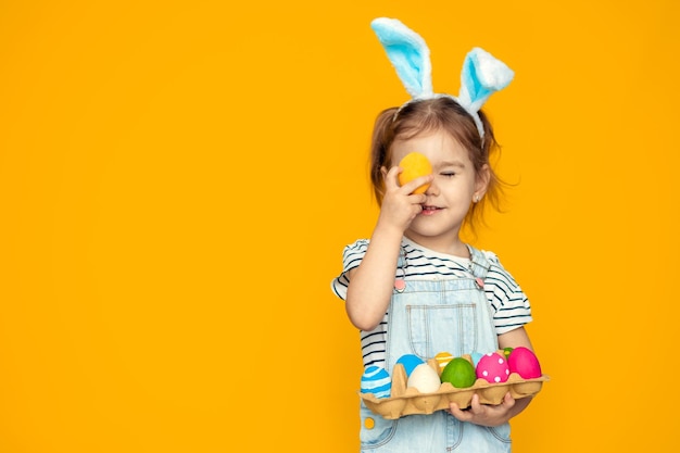 Funny happy baby girl with easter eggs and bunny ears on yellow background