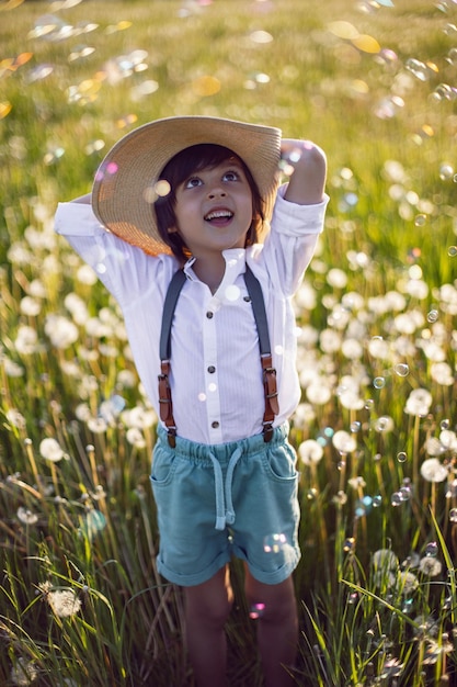 Funny happy a beautiful boy child in a hat stands on a field with white dandelions at sunset in summer soap bubbles are flying