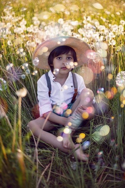 Funny happy a beautiful boy child sit in hat on a field with white dandelions at sunset in summer soap bubbles are flying