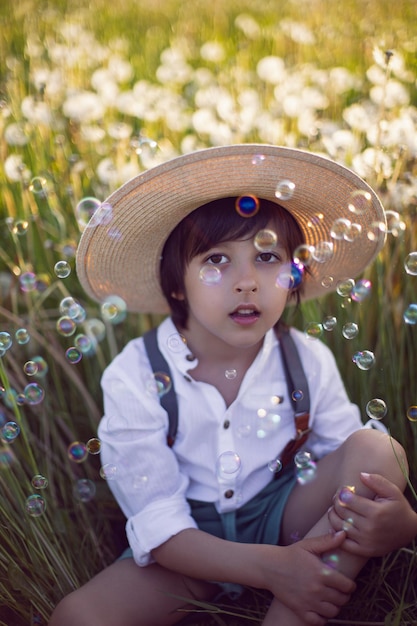 Funny happy a beautiful boy child sit in hat on a field with white dandelions at sunset in summer soap bubbles are flying