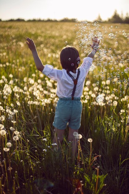 웃긴 행복한 아름다운 소년 아이는 여름 비누방울이 날고 있는 일몰에 흰 민들레가 있는 들판에 서 있다