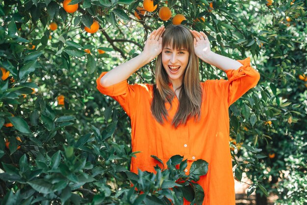 La ragazza felice divertente in vestito arancione sta posando alla macchina fotografica mettendo le mani sulla testa nel giardino degli aranci