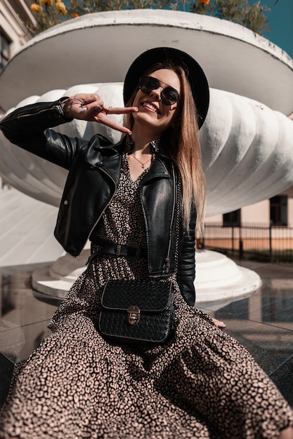 funny hipster girl with sunglasses and a hat in a stylish dress with a fashionable handbag and leather jacket shows the peace sign and sits in the city