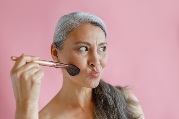 Funny hoary haired Asian lady applies makeup with brush grimacing on pink background