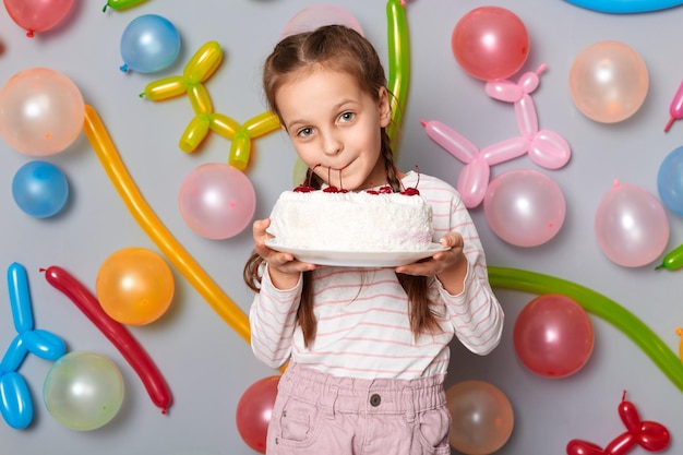 Funny hungry little girl with pigtails standing against gray wall decorated with colorful balloons holding licking cake at her birthday party