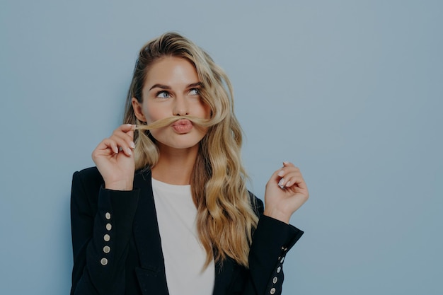 Funny joyful blonde woman having fun while posing against blue studio wall playing with strand of hair