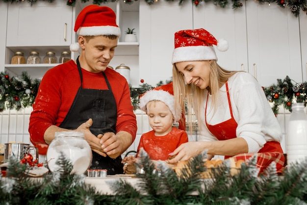 クリスマスの帽子と小麦粉で汚れた服を着たおかしなうれしそうな家族が、キッチンのテーブルに立って生地をこねている