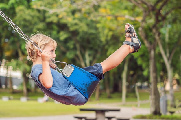 Funny kid boy having fun with chain swing on outdoor playground. child swinging on warm day. Active leisure with kids. Boy wearing casual colorful school kid clothes