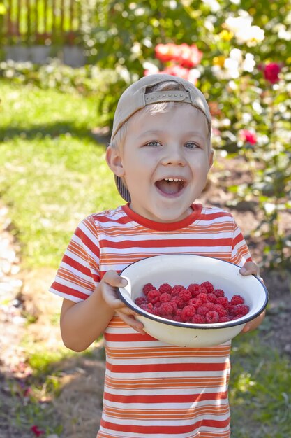 Funny kid boy with bowl of a ripe raspberry outdoor