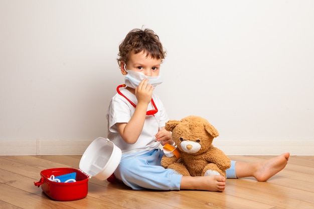 Funny kid in a medical mask plays with a teddy bear at the doctor.