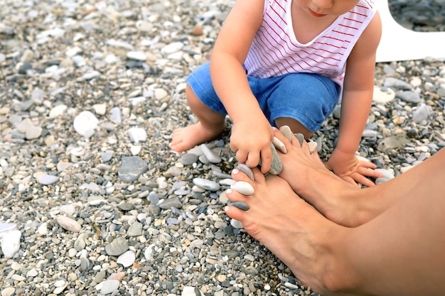 자갈 해변에서 재미있는 아이는 그의 어머니와 함께 그의 손가락 해양 개념 사이에 돌을 삽입합니다