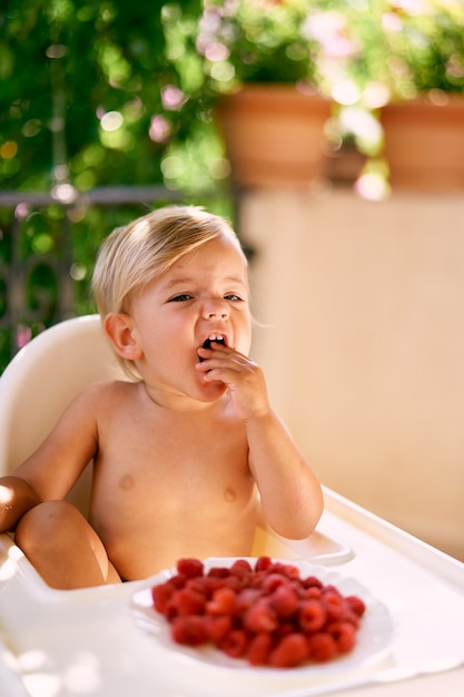 Funny kid sits on a high chair on the balcony and eats raspberries