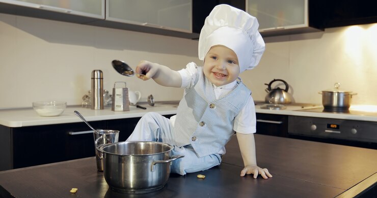 Photo a funny kid with the fork and spoon in the kitchen portrait of a dirty boy in a culinary hat