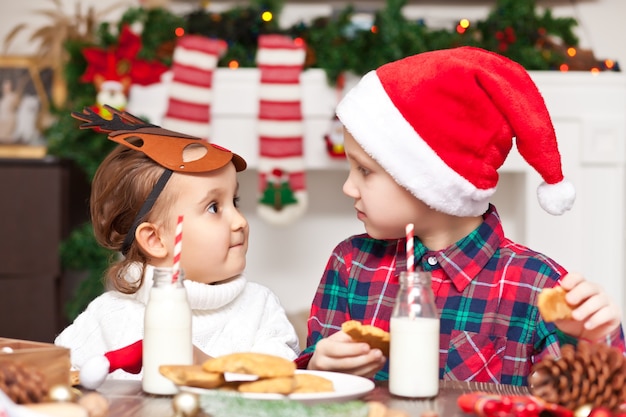Смешные дети девочка и мальчик в кепке Санты пьют рождественское молоко, едят печенье и используют портативный компьютер с цифровым планшетом. Звоните по сети друзьям или родителям и получайте удовольствие.