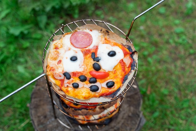 Funny kids homemade pizza with face cheese eyes black olives\
tomato mouth sausage sauce family outdoor cooking in oriental oven\
tandoor special metal rack for barbecue vegetables green grass