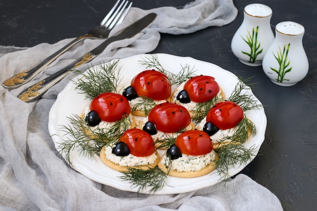 Забавная закуска в форме божьей коровки с помидорами на крекерах с сыром на белой тарелке