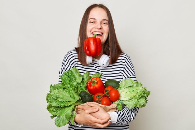 Funny laughing brown haired young woman embraces bouquet of fresh vegetables wearing striped casual shirt holding red pepper in mouth posing isolated over gray background