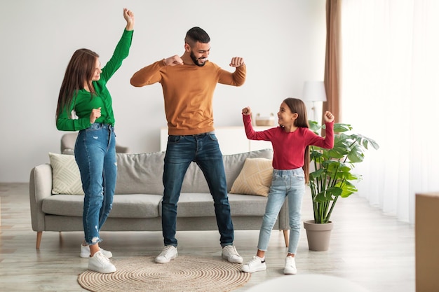 面白い余暇活動の概念。陽気なアラブの父は彼の小さな子供と妻と一緒に音楽に移動して踊っています。居間で家族との自由な時間を楽しんで楽しんでいる男、女、女
