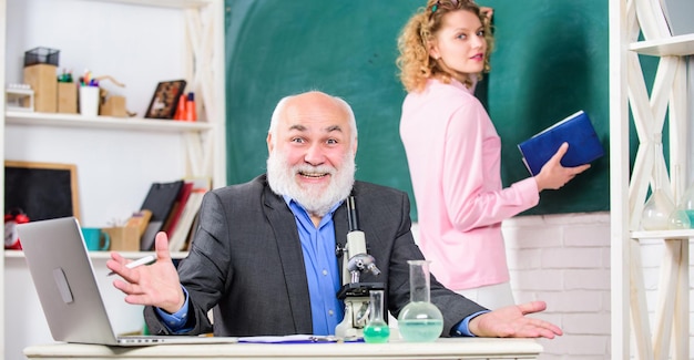 Foto lezione divertente insegnante maturo con bicchiere e microscopio torna a scuola studentessa ragazza al consiglio studente e tutor con laptop lezione di biologia scienza e biotecnologia ricerca educazione chimica