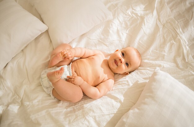 Забавная маленькая девочка в подгузнике, лежа на кровати и держась за ноги руками