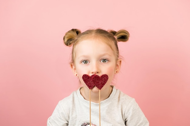 Funny little blonde girl smiling and playing with red heart toy on pink background. Valentines Day or Mothers Day concept.
