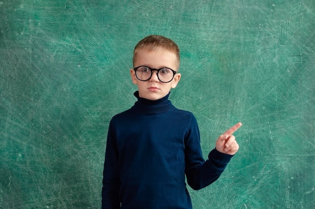 Забавный маленький мальчик в синем свитере и очках из начальной школы на фоне доски. Ребенок проходит проверку зрения.