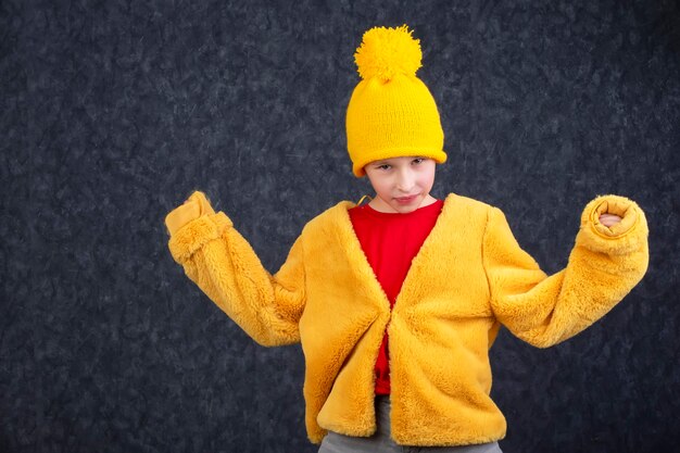 Photo funny little boy in a fluffy yellow fur coat and a knitted hat on a gray background.