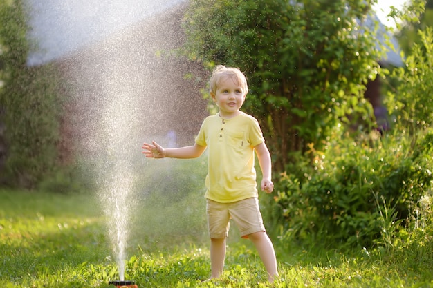 Funny little boy playing with garden sprinkler in sunny backyard