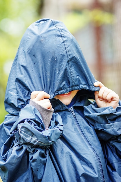 面白い小さな男の子は、屋外のフード付きのレインコートで雨の中を歩きます