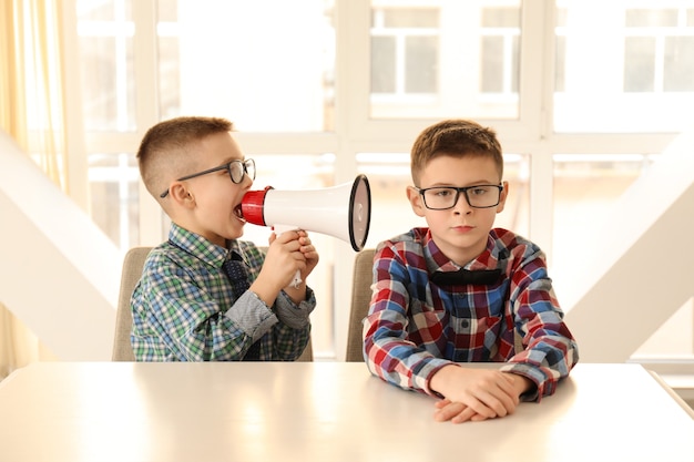 Funny little boy with megaphone shouting at his bored friend sitting at table