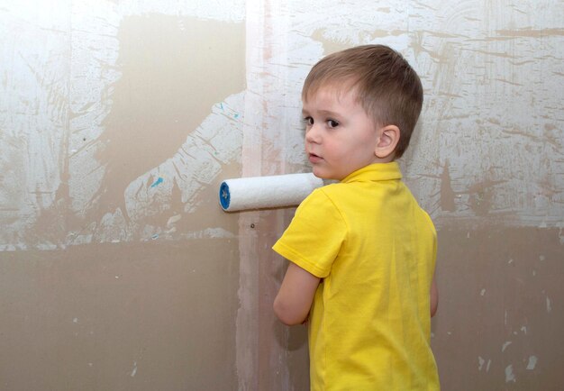 Funny little boy in yellow shirt paints wall with paint roller in new house. Family apartment renovation.