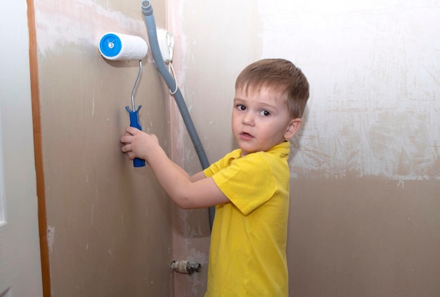 Funny little boy in yellow shirt paints wall with paint roller\
in new house. family apartment renovation.
