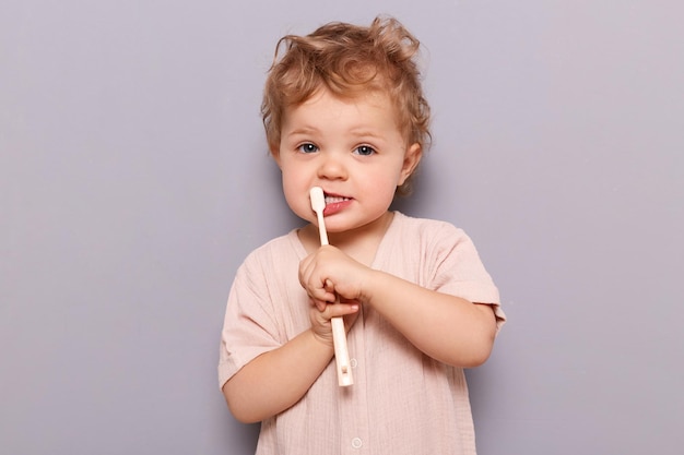 Funny little child baby brushing teeth isolated over gray background oral care using kid's toothbrush looking at camera wearing tshirt