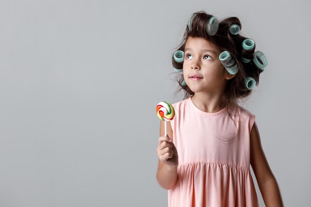Funny little child girl in pink dress and hair curlers holding lollipop on gray background.